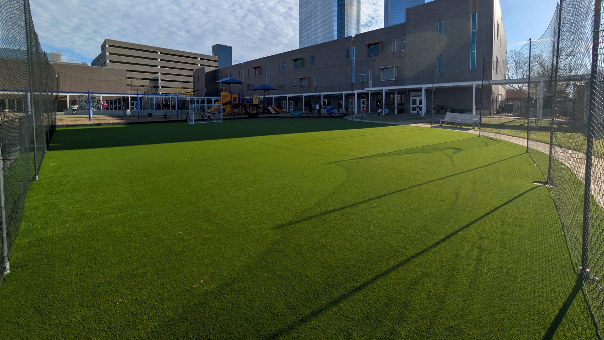 Large artificial turf play field inside a netted enclosure, with a school playground and the downtown Oklahoma City skyline behind it