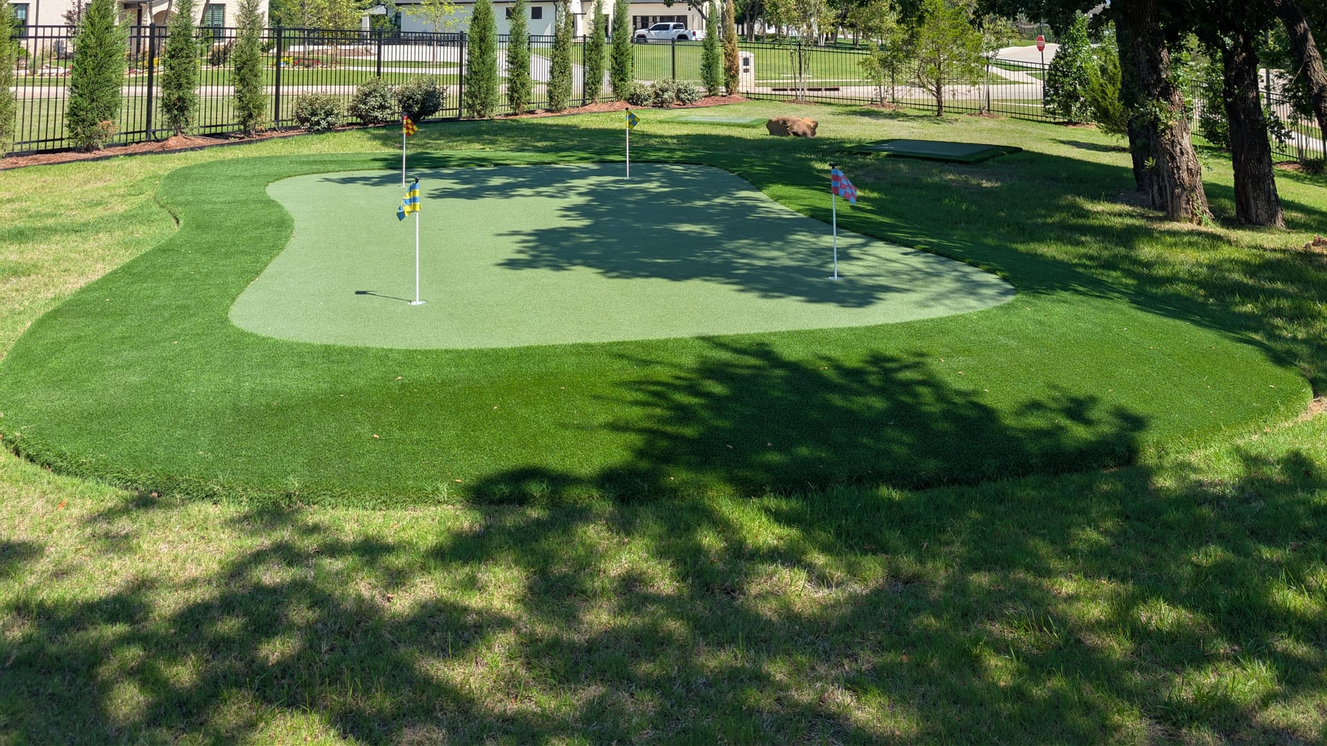 Backyard artificial putting green with two flagged cups, installed under a mature shade tree beside a wrought iron fence